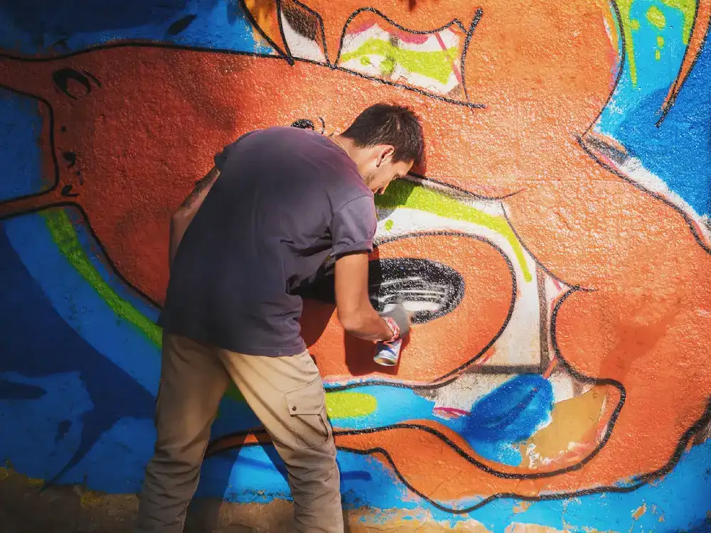 Artist using spray paint to create a vibrant, large-scale orange and blue mural on an outdoor wall.