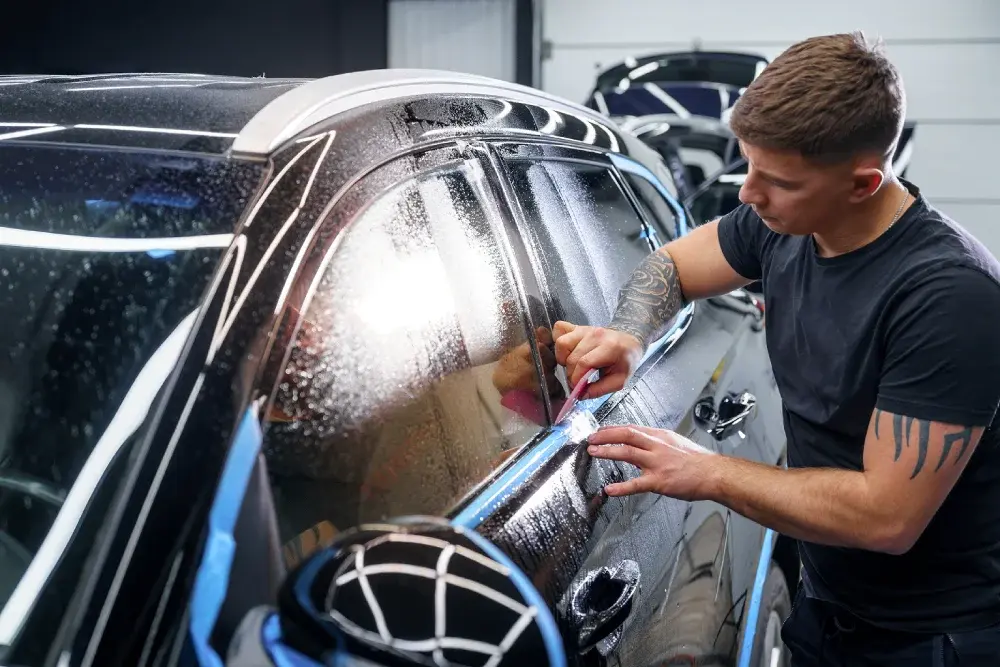 A professional installer using a squeegee to apply and smooth window tint film on a black vehicle at Katazoom.